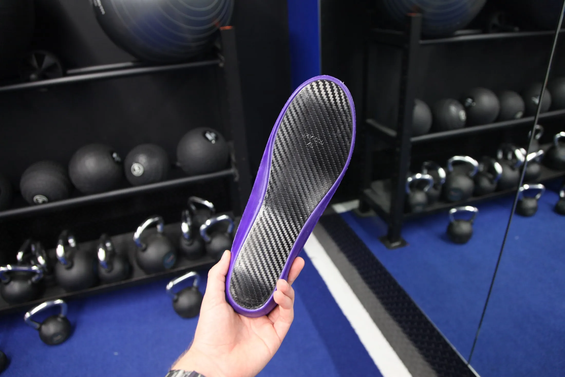 Hand holding a CRBN V2 up in the gym, carbon fibre underside facing the camera