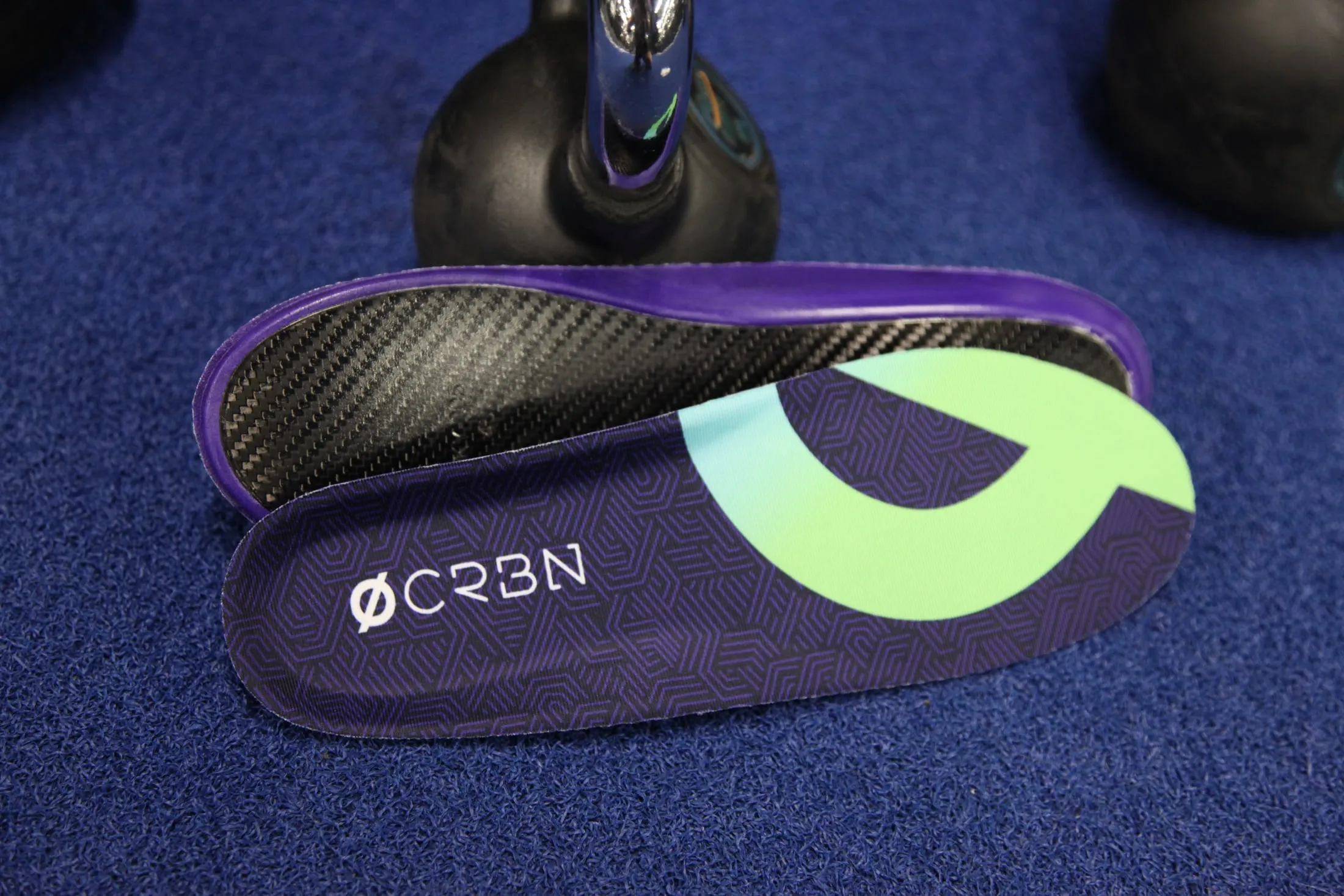 A CRBN V2 pair on blue gym carpet beside a kettlebell, carbon underside showing